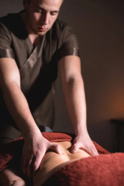 A male professional massage therapist does back massage to a female client in a dark cozy medical room against a background of burning candles. Low key. — Stock Photo, Image