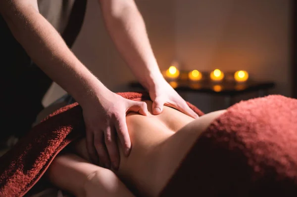 A male professional massage therapist does back massage to a female client in a dark cozy medical room against a background of burning candles. Low key. — Stock Photo, Image