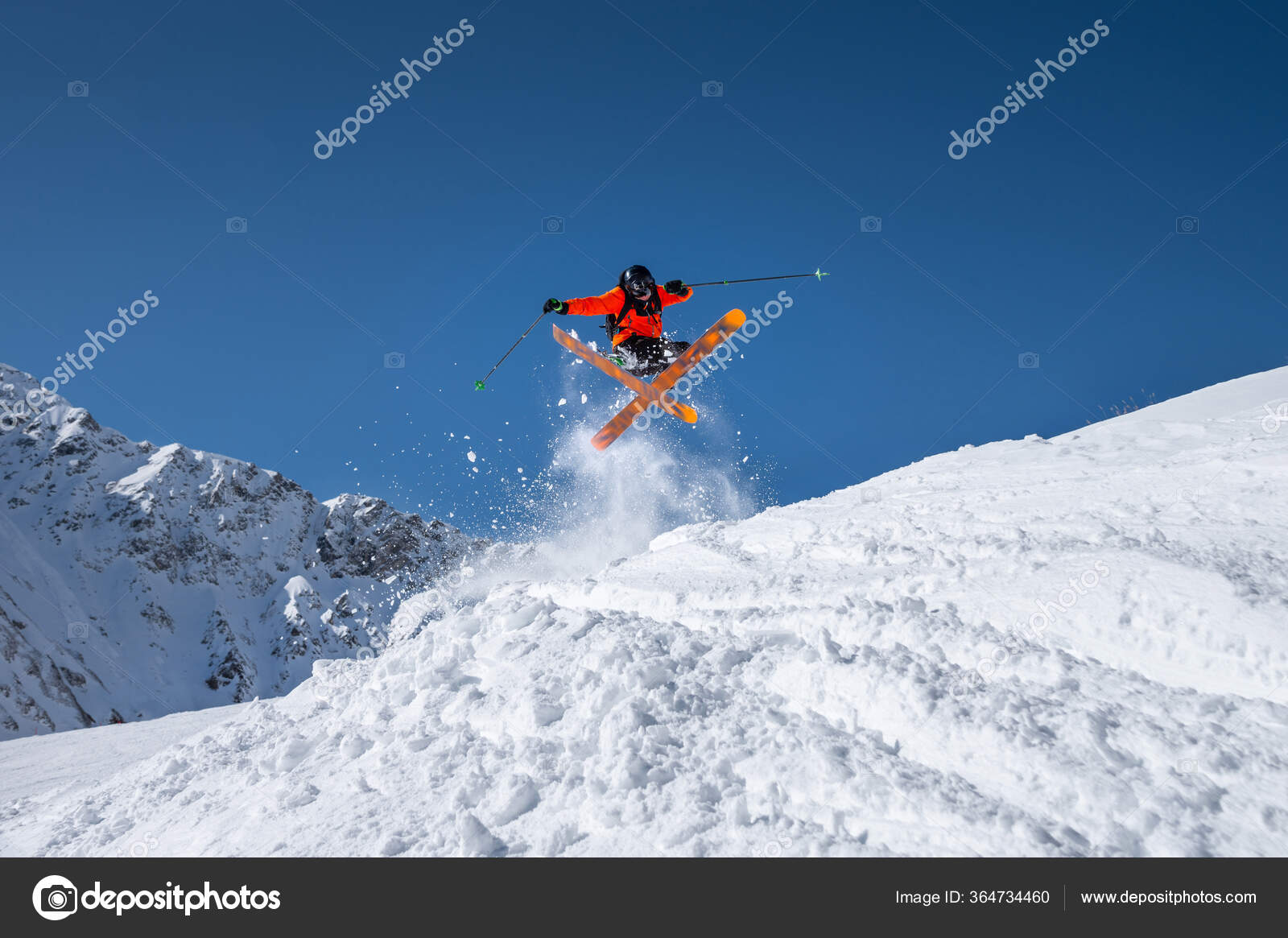 Male athlete skier in an orange trigger makes a jump trick with flying snow  powder against the backdrop of snow-capped Caucasus mountains and a blue  sky. Winter Extreme Sports Concept Stock Photo