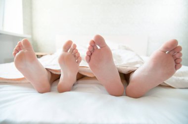 Yatak odasındaki yorganın altından dışarı bakan genç bir çiftin çıplak ayakları. Salgın sırasında kendini izole eden sağlıklı ayak derisi kavramı.
