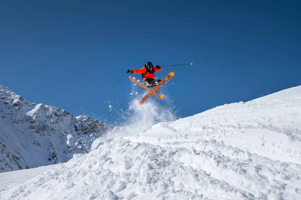 O atleta masculino esquiador em um gatilho laranja faz um truque de salto com pó de neve voador contra o pano de fundo das montanhas do Cáucaso cobertas de neve e um céu azul. Inverno conceito de esportes radicais — Fotografia de Stock