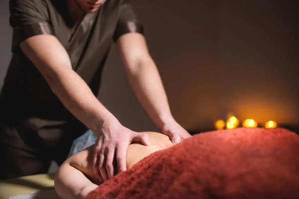 A male professional massage therapist does back massage to a female client in a dark cozy medical room against a background of burning candles. Low key.