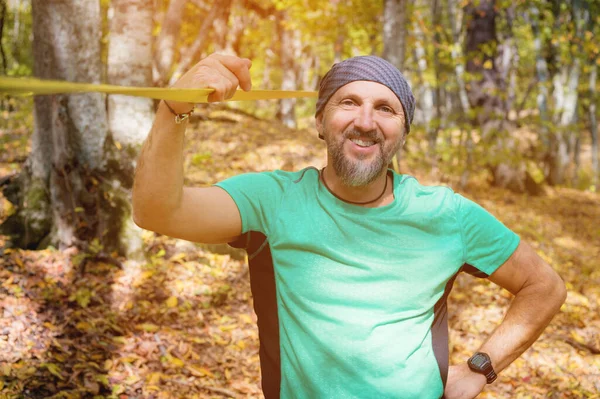 Retrato de um homem atlético barbudo envolvido em folga ao lado de uma funda esticada para equilíbrio na floresta de outono. Atleta sorridente 40 anos em um dia ensolarado — Fotografia de Stock