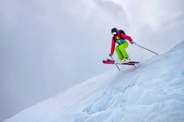 Ein Mädchen im grünen Skianzug springt bei trübem Wetter von einem schneebedeckten Felsvorsprung gegen einen bewölkten Himmel. Sonne scheint durch die Wolken. Das Konzept von Wintersport und Extremsport — Stockfoto