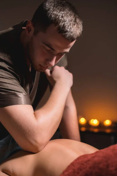 A professional male masseur makes an massage to a female client with an elbow against the background of burning candles in a dark room of a massage room. Alternative Massage Concept — Stock Photo, Image