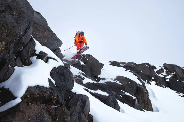 Άντρας αθλητής σκιέρ σε πορτοκαλί σκανδάλη με γκρι σακίδιο και κοντάρια σκι πηδώντας από μια ψηλή βραχώδη χιονισμένη πλαγιά με φόντο τα βουνά στα σύννεφα σε μια συννεφιασμένη μέρα. — Φωτογραφία Αρχείου