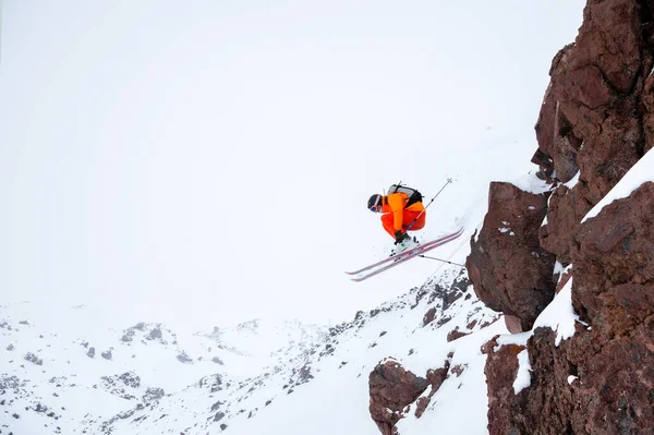 Mannelijke atleet skiër in een oranje trekker met een grijze rugzak en skistokken springen van een hoge rotsachtige besneeuwde helling tegen de achtergrond van bergen in de wolken op een bewolkte dag. — Stockfoto