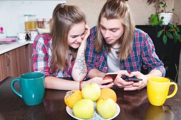 An attractive young Caucasian couple in the kitchen at the dinner table drinks tea and uses their smartphones. — Stock Photo, Image