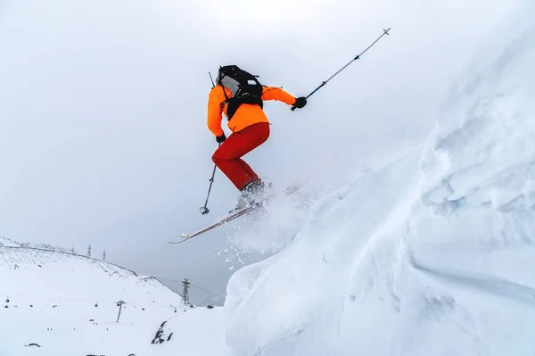 Άντρας αθλητής σκιέρ σε πορτοκαλί σκανδάλη με γκρι σακίδιο και κοντάρια σκι πηδά από μια ψηλή χιονισμένη πλαγιά με φόντο τα βουνά στα σύννεφα σε μια συννεφιασμένη μέρα. — Φωτογραφία Αρχείου