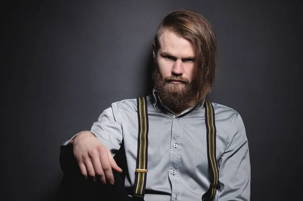 Contrastando retrato de bajo perfil de un hombre barbudo de pelo largo con estilo brutal en una camisa gris con tirantes mira severamente a la cámara. El concepto de estilo y belleza brutal en el estudio — Foto de Stock