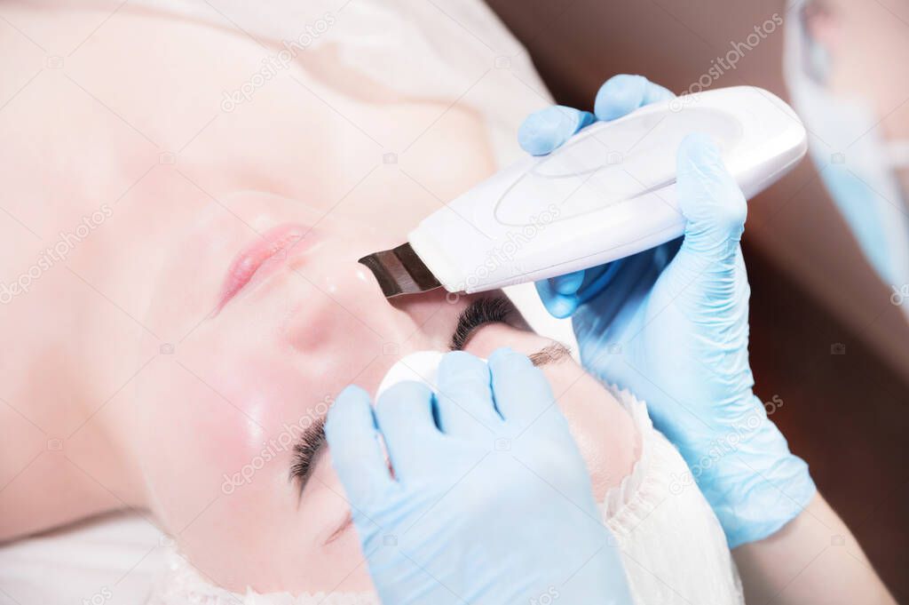 Close-up ultrasound face peel at the beautician. Beautician squeezes acne on the patients forehead with a medical needle. Face next to hands in blue gloves of cosmologist