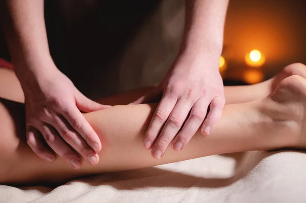 Närbild professionell kalv muskel massage till en kvinnlig klient av en manlig sjukgymnast i en massage salong i ett mörkt kontor mot bakgrund av brinnande ljus — Stockfoto