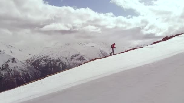 Воздушный молодой человек с длинными волосами в солнцезащитных очках и в rbczak поднимается на скалистый хребет с отслеживающими палками высоко в горах рядом со снежной сменой. вид сбоку — стоковое видео