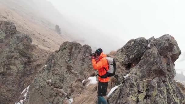 Letecký pohled na mladého fotografa s fotoaparátem v ruce stojí na vysoké skále v rokli a fotografuje alpský vodopád během sněhové bouře a sněžení na jaře. — Stock video
