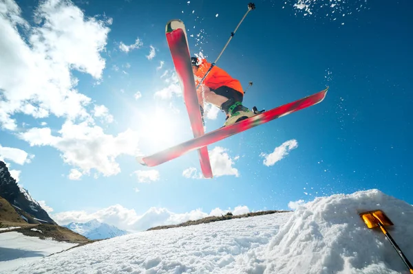 Um jovem atleta esquiador faz um truque em um salto de kicker de neve e esquis cruzados em uma luz de contador contra um céu azul e nuvens nas montanhas . — Fotografia de Stock