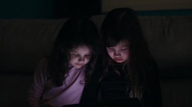 Genç kız kardeşler evlerinde kanepede, karanlıkta elektronik aletlerle.