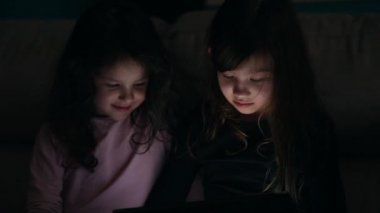 Genç kız kardeşler evlerinde kanepede, karanlıkta elektronik aletlerle.
