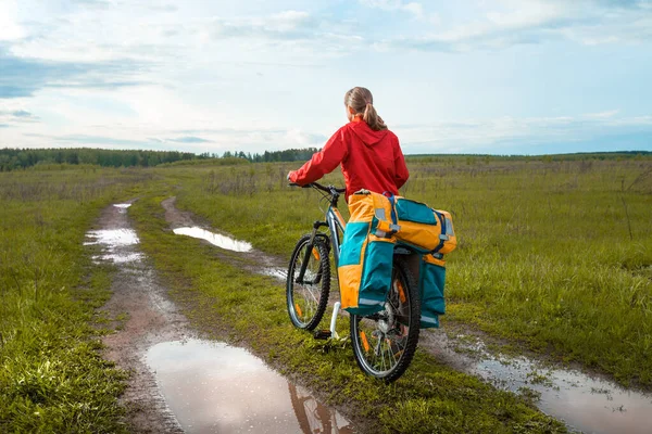 年轻的骑自行车的女徒步旅行者站在草地上潮湿的乡间路上 — 图库照片