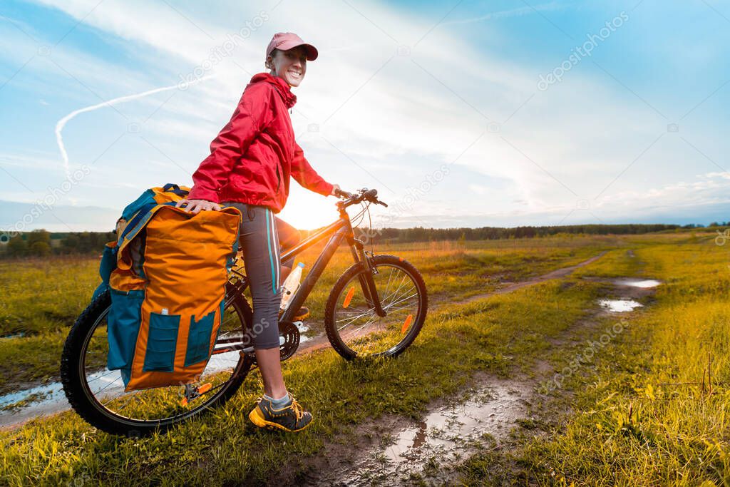 Young lady hiker with loaded bicycle standing on a wet rural road in the meadow