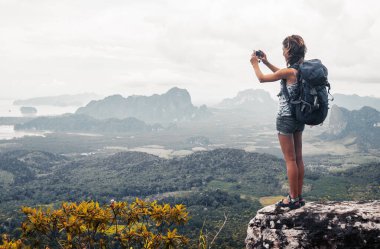 Sırt çantalı genç bayan yürüyüşçü dağın tepesinde duruyor ve vadide fotoğraf çekiyor.