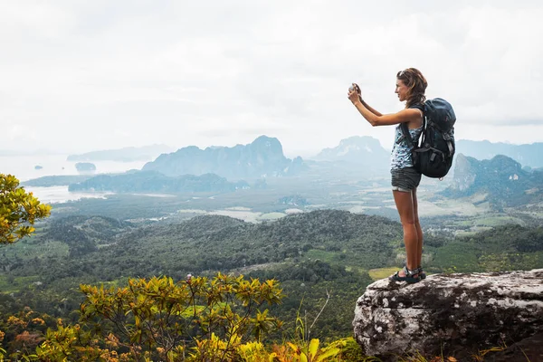 年轻的女徒步旅行者背着背包站在山顶上给山谷拍照 — 图库照片