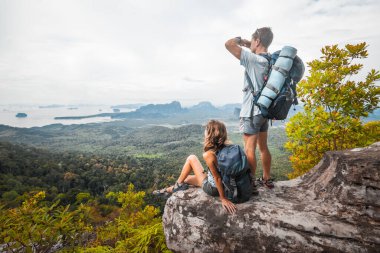 Sırt çantalı iki turist dağın tepesinde dinleniyor ve vadi manzarasının tadını çıkarıyorlar.