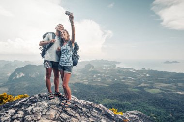 İki yürüyüşçü arka planda vadi manzaralı dağın tepesinden selfie çekiyor.