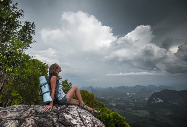 Sırt çantalı genç bir kadın uçurumda oturuyor ve vadi manzarasının tadını çıkarıyor.