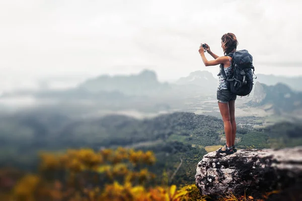 年轻的女徒步旅行者背着背包站在山顶上给山谷拍照 — 图库照片