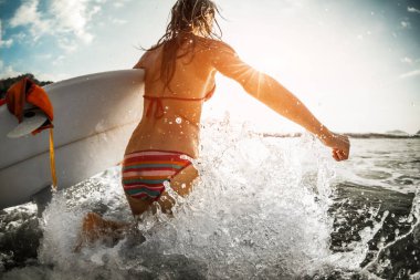 Kadın elinde sörf tahtası ve bir sürü su sıçramasıyla okyanusa koşuyor.