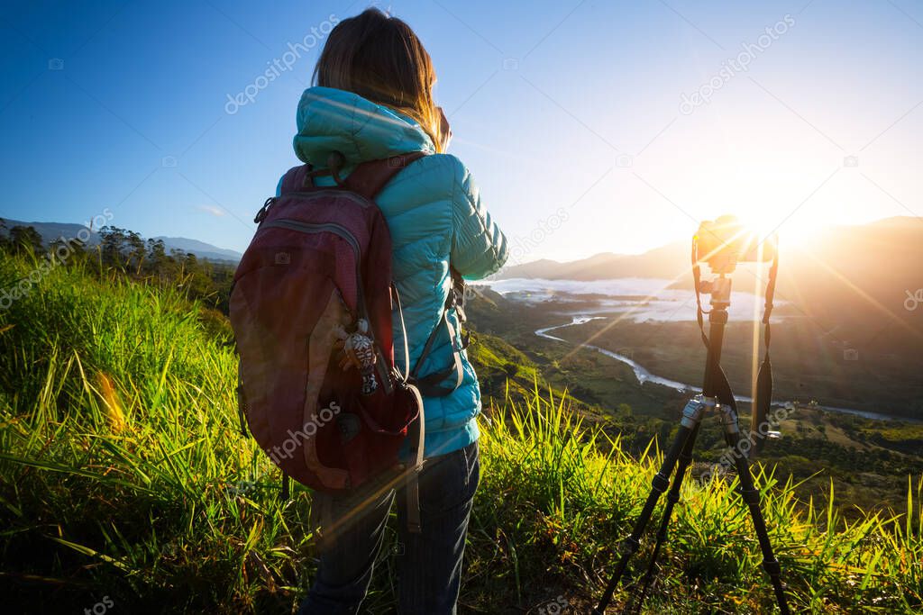 Photographer stans on the meadow with camera set on tripod during sunrise