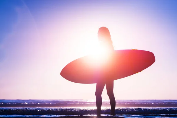 Woman with surfboard walks on the beach during sunset