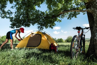 Genç bayan bir çayırda çadır kuruyor, ağacın altında yeşil çimenler var.