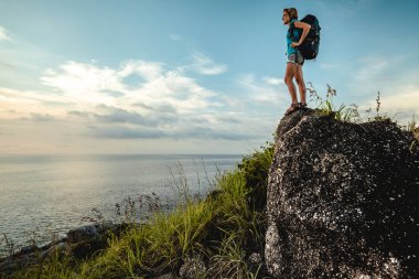 Sırt çantalı bayan yürüyüşçü taşın üzerinde duruyor ve deniz manzarasının tadını çıkarıyor.
