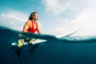 Genç sörfçü güneşli bir günde dalgayı okyanusta bekler. Görüntüyü su altı görüntüsüyle böl