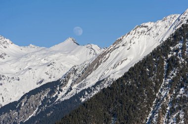 Panoramik manzara karla kaplı dağ sırasındaki vadide kozalaklı çam ağaçları ve mavi gökyüzü arka planında ay var.