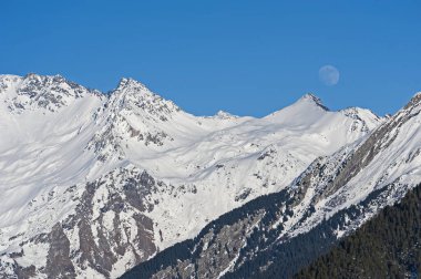 Panoramik manzara karla kaplı dağ sırasındaki vadide kozalaklı çam ağaçları ve mavi gökyüzü arka planında ay var.