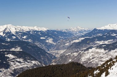 Karla kaplı bir dağ sırasının panoramik manzarası, yamaç paraşütçüleriyle vadiden aşağı bakıyor.