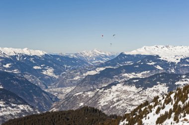 Karla kaplı bir dağ sırasının panoramik manzarası, yamaç paraşütçüleriyle vadiden aşağı bakıyor.