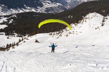 Paraglider karla kaplı bir yamaçtan havalanıyor ormanlı bir dağ vadisine bakıyor.