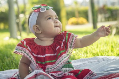 Bebita blanca muy hermosa, sentada en la hierba, con vestido de colores mexicanos, cinta blanca y lazo de colorines en la cabeza. Gesticulando y expresando emociones diversas, originales y muy simpticas, graciosas, sugerentes. Çok doğal sonbahar.