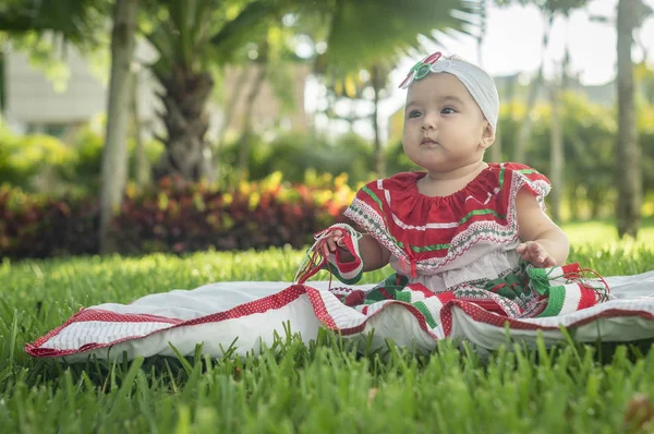 Bebê branco muito bonito, sentado na grama, vestindo vestido colorido mexicano, fita branca e gravata borboleta na cabeça. Gesticulando e expressando emoções diversas, originais e muito agradáveis, engraçadas, sugestivas. Muito natural e autêntico. Sol brilhante — Fotografia de Stock