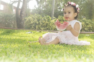 Güzel, beyaz çiçek kafasındaki bir Bahçe, yeşil çimenlerin üzerine oturmuş kafa bandı ile bir dilim o onun elinde tutan karpuz yeme kız