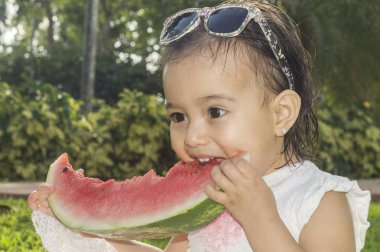 İki-yıl-yaşlı kız, beyaz ve güzel, karpuz ya da elinde tutar kırmızı kavun yemek başına, elbise, yeşil çimenlerin üzerinde oturan, kafa bandı çiçek pembe. Komik, komik ifadeler gülümseyen iskambil
