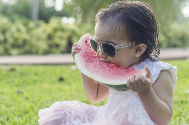 İki-yıl-yaşlı kız, beyaz ve güzel, karpuz ya da elinde tutar kırmızı kavun yemek başına, elbise, yeşil çimenlerin üzerinde oturan, kafa bandı çiçek pembe. Komik, komik ifadeler gülümseyen iskambil