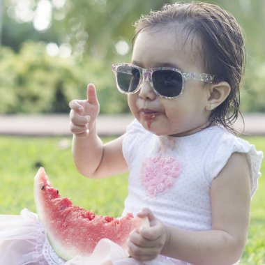 İki-yıl-yaşlı kız, beyaz ve güzel, karpuz ya da elinde tutar kırmızı kavun yemek başına, elbise, yeşil çimenlerin üzerinde oturan, kafa bandı çiçek pembe. Komik, komik ifadeler gülümseyen iskambil