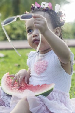 İki-yıl-yaşlı kız, beyaz ve güzel, karpuz ya da elinde tutar kırmızı kavun yemek başına, elbise, yeşil çimenlerin üzerinde oturan, kafa bandı çiçek pembe. Komik, komik ifadeler gülümseyen iskambil