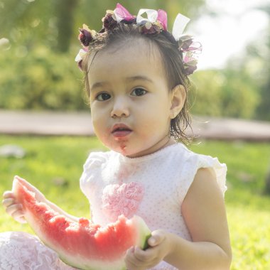 İki-yıl-yaşlı kız, beyaz ve güzel, karpuz ya da elinde tutar kırmızı kavun yemek başına, elbise, yeşil çimenlerin üzerinde oturan, kafa bandı çiçek pembe. Komik, komik ifadeler gülümseyen iskambil