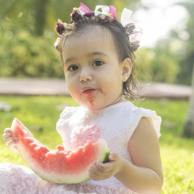 İki-yıl-yaşlı kız, beyaz ve güzel, karpuz ya da elinde tutar kırmızı kavun yemek başına, elbise, yeşil çimenlerin üzerinde oturan, kafa bandı çiçek pembe. Komik, komik ifadeler gülümseyen iskambil
