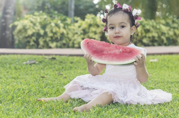 Menina branca e bonita, com uma faixa de flores na cabeça, sentada no relvado verde de um jardim, a comer uma fatia de melancia que ela segura nas mãos. — Fotografia de Stock